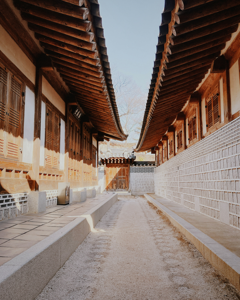 Narrow stone alleyway lined with traditional hanok houses in a historic Korean village, featuring wooden doors and tiled roofs.