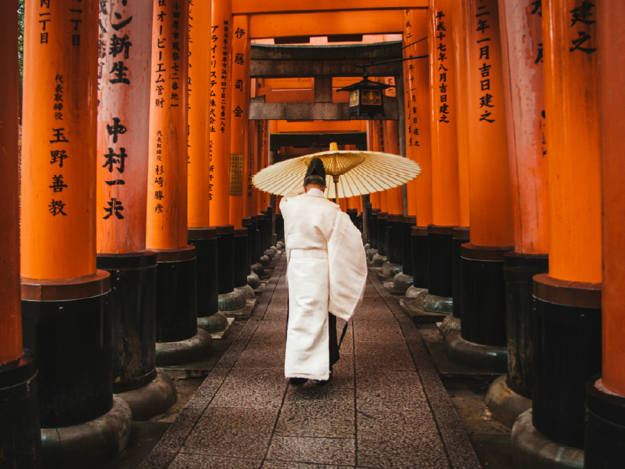 A person wearing traditional Japanese attire and holding a parasol walks through a tunnel of vibrant orange torii gates, Fushimi Inari Shrine, Kyoto, Japan