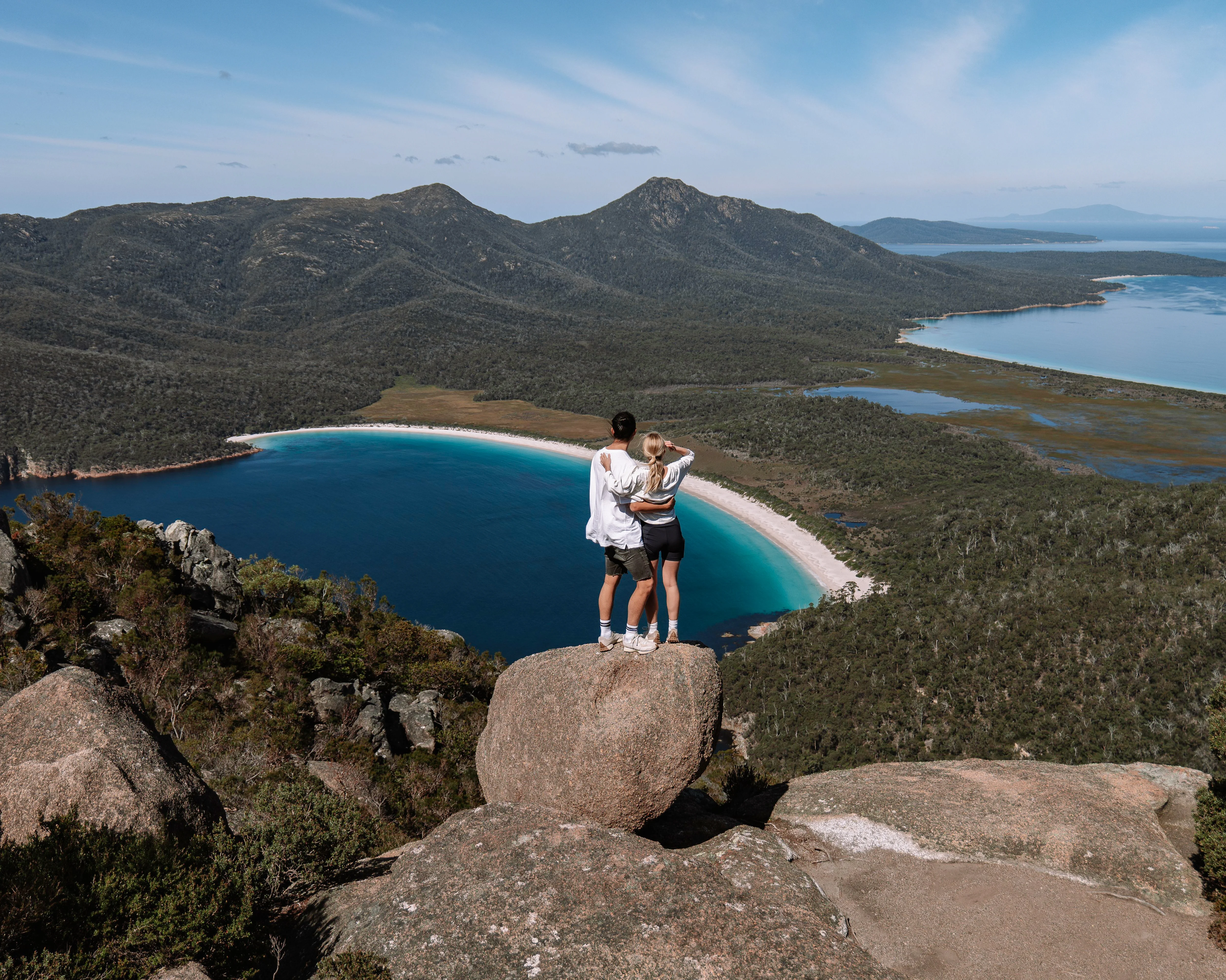 Two hikers on Mount Amos summit in Freycinet National Park Tasmania overlooking Wineglass Bay and Tasman Sea under clear blue sky