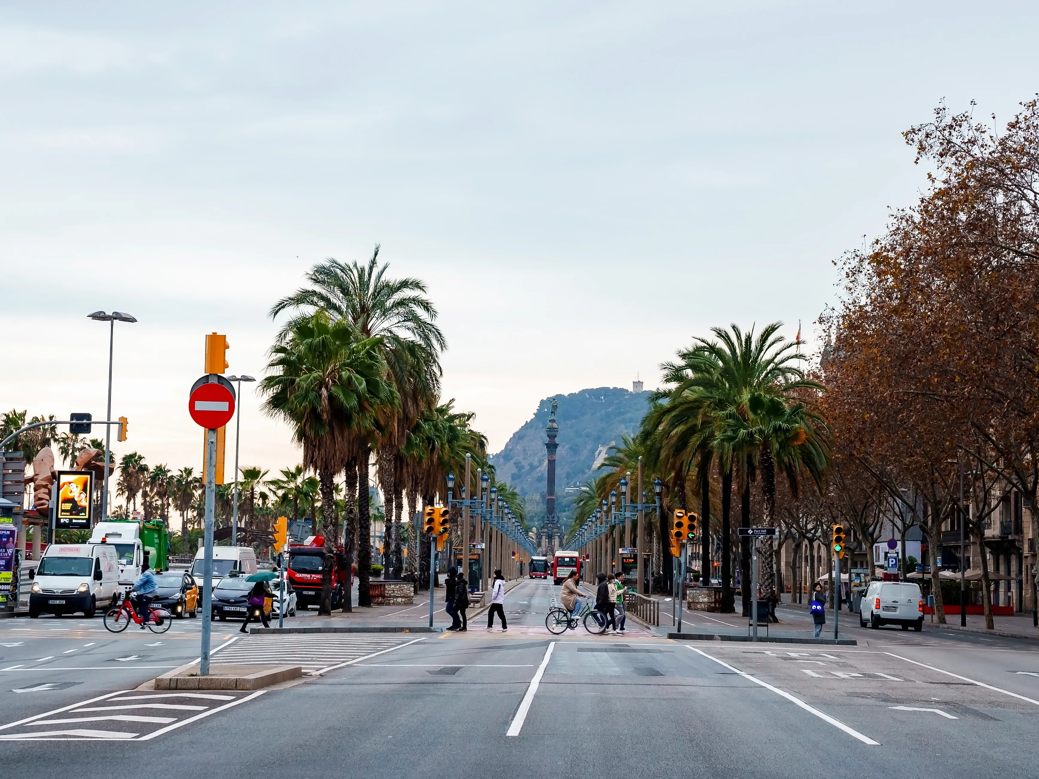 La Rambla in Barcelona, Spain, showing a wide palm‑lined boulevard with pedestrians crossing, cyclists, traffic lights, and the city street stretching toward the hills.