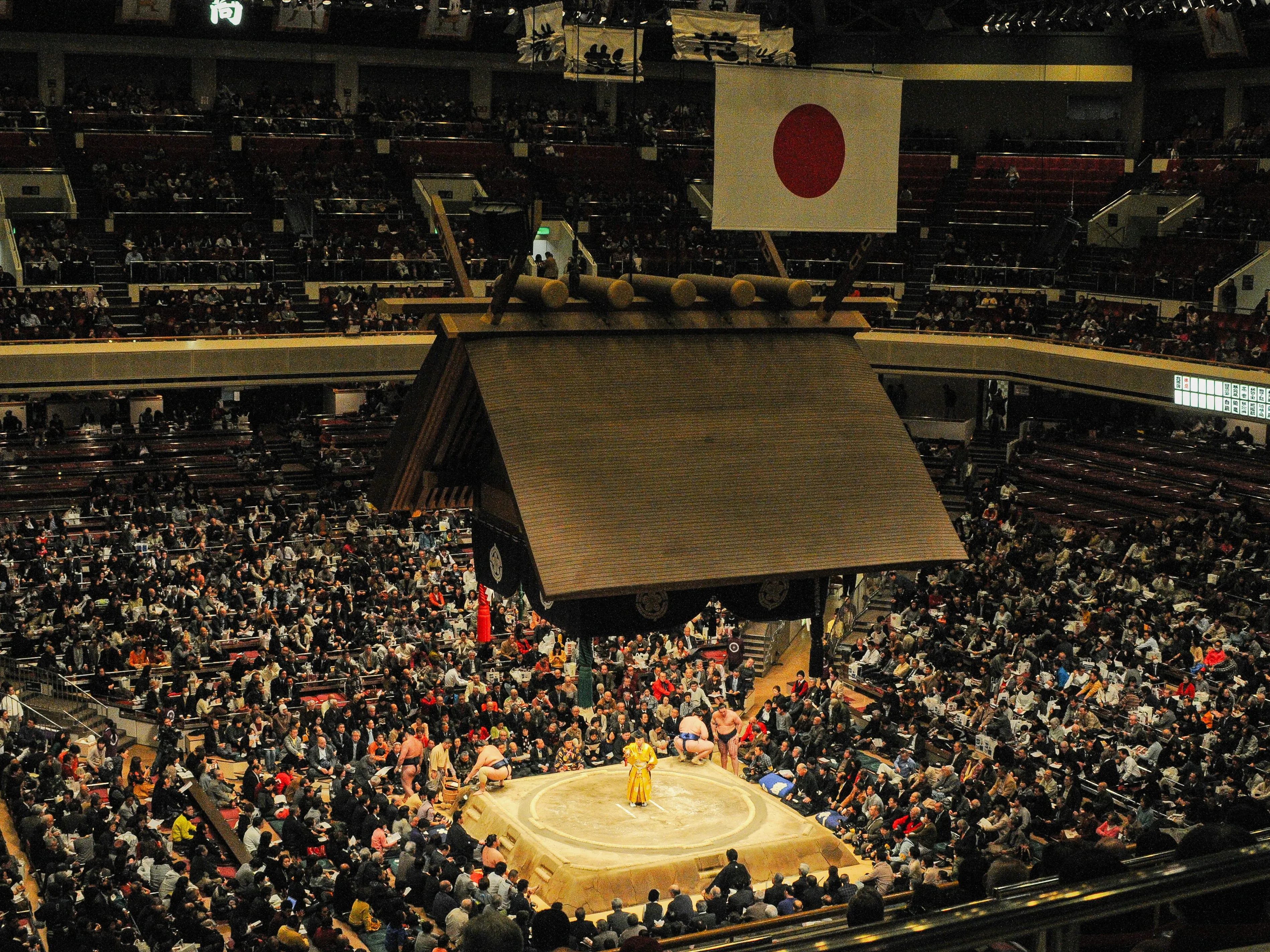 Sumo wrestlers competing inside a traditional stadium with spectators and the Japanese flag overhead.