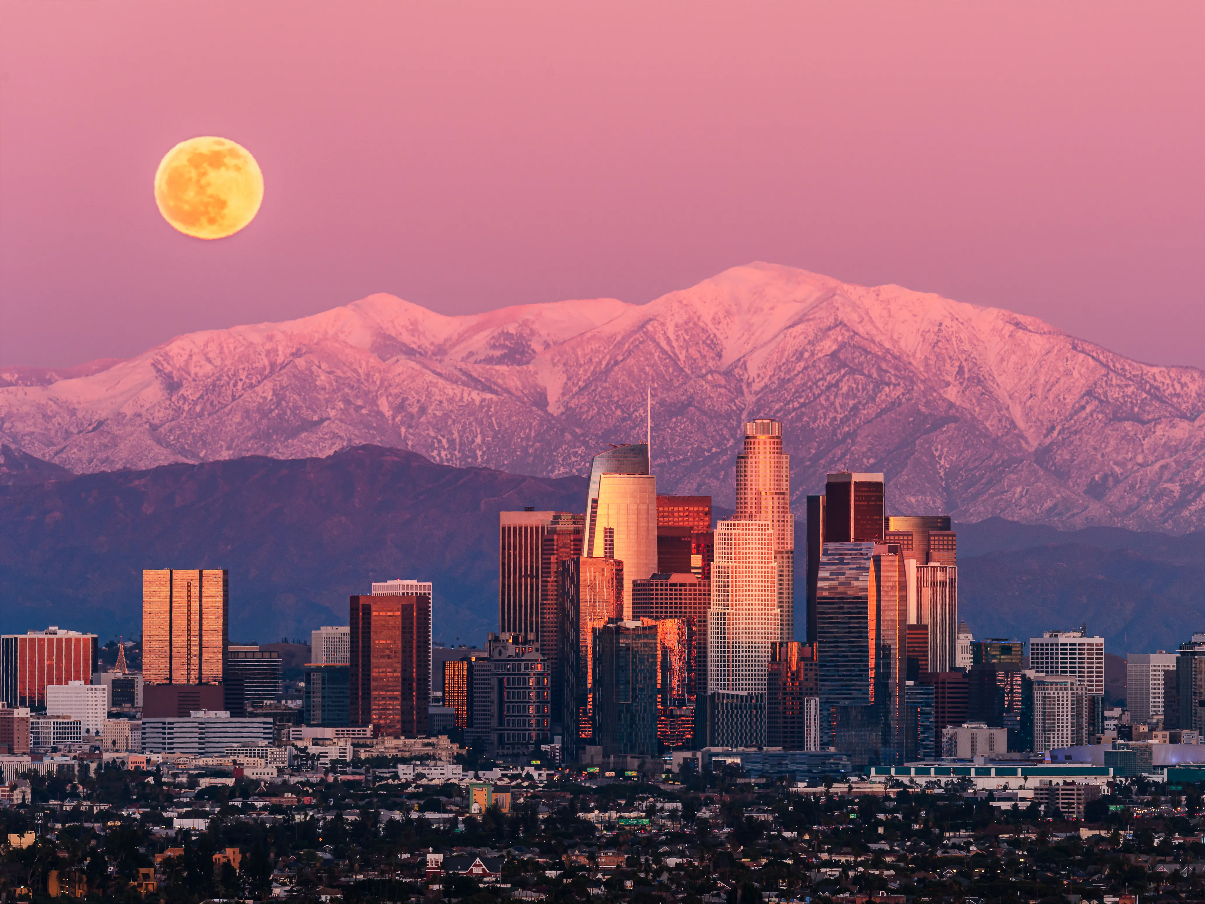 Los Angeles downtown skyline at sunset with a full moon rising over snow-capped San Gabriel Mountains — a direct flight destination from Australia.
