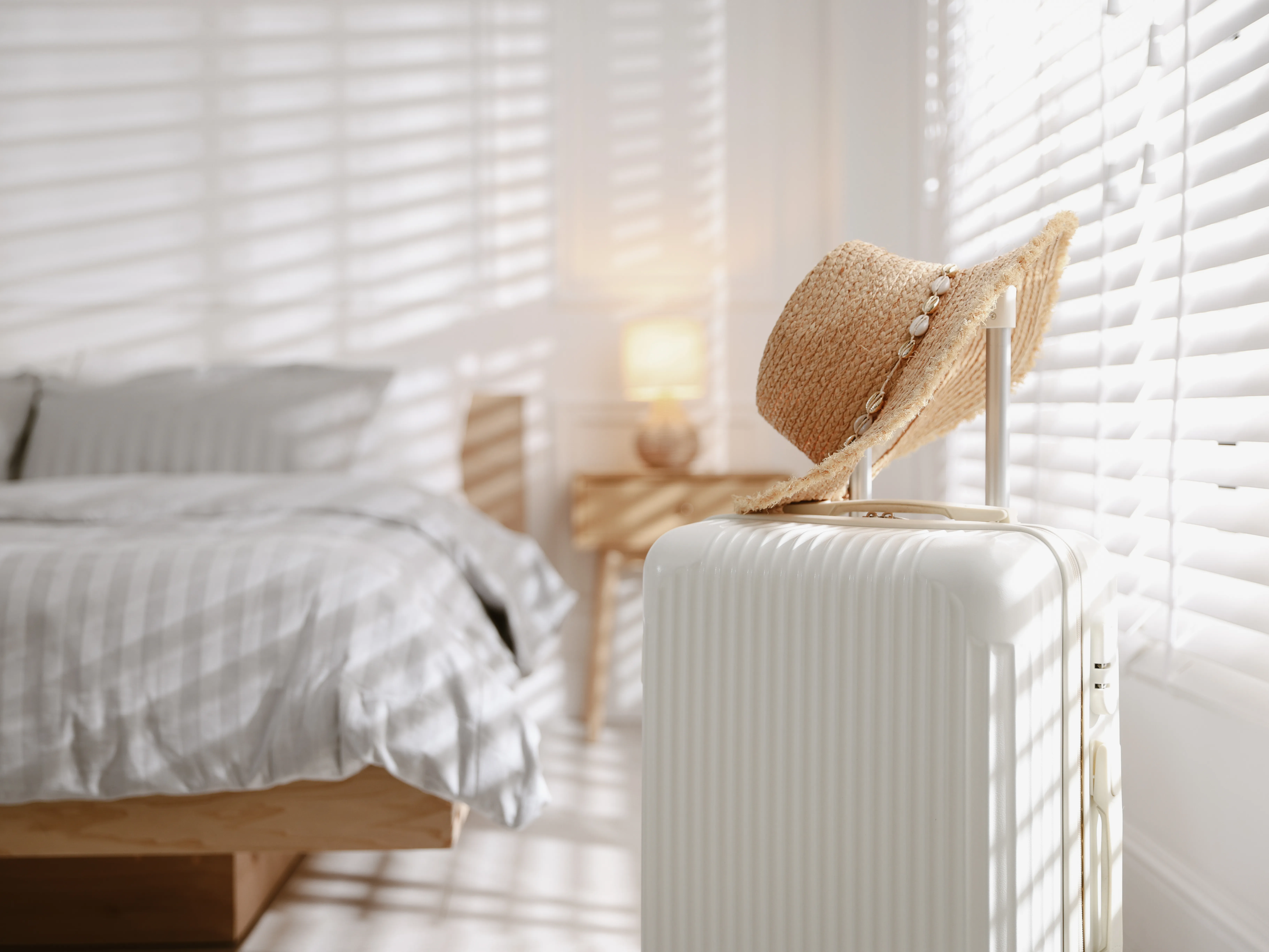 A compact white carry-on suitcase with a straw hat resting on the handle, standing in a bright bedroom before a trip.