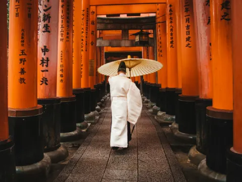 A person wearing traditional Japanese attire and holding a parasol walks through a tunnel of vibrant orange torii gates, Fushimi Inari Shrine, Kyoto