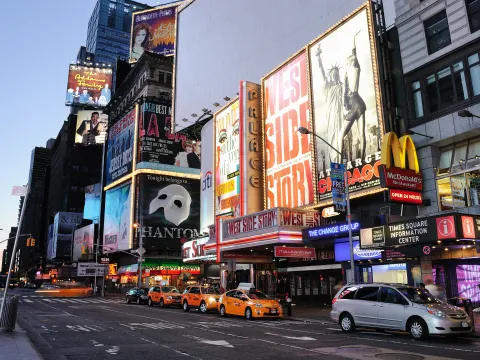 Times Square in New York City at dusk, showing illuminated Broadway theatre billboards, yellow taxis, and busy city streets.